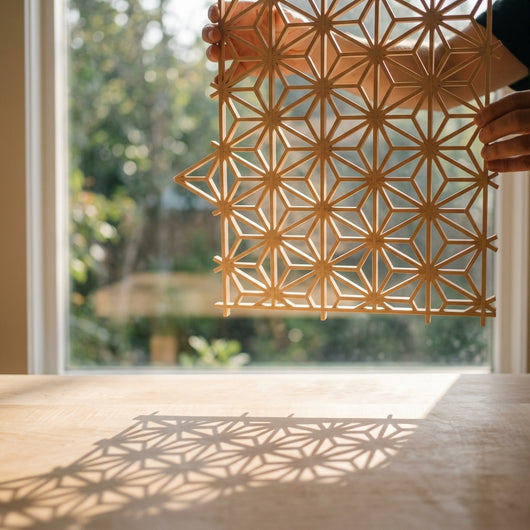 Finished kumiko lattice panel held up to window light, casting geometric asa-no-ha shadows on maple surface